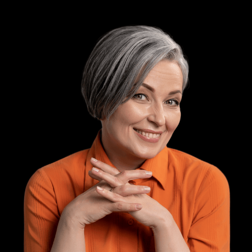 Smiling mature woman with short gray hair in an orange blouse, hands folded, exuding warmth and confidence on a black background