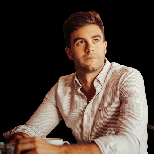 Confident young man in a striped shirt sitting at a table, looking thoughtful with a slight smile, isolated on a black background.